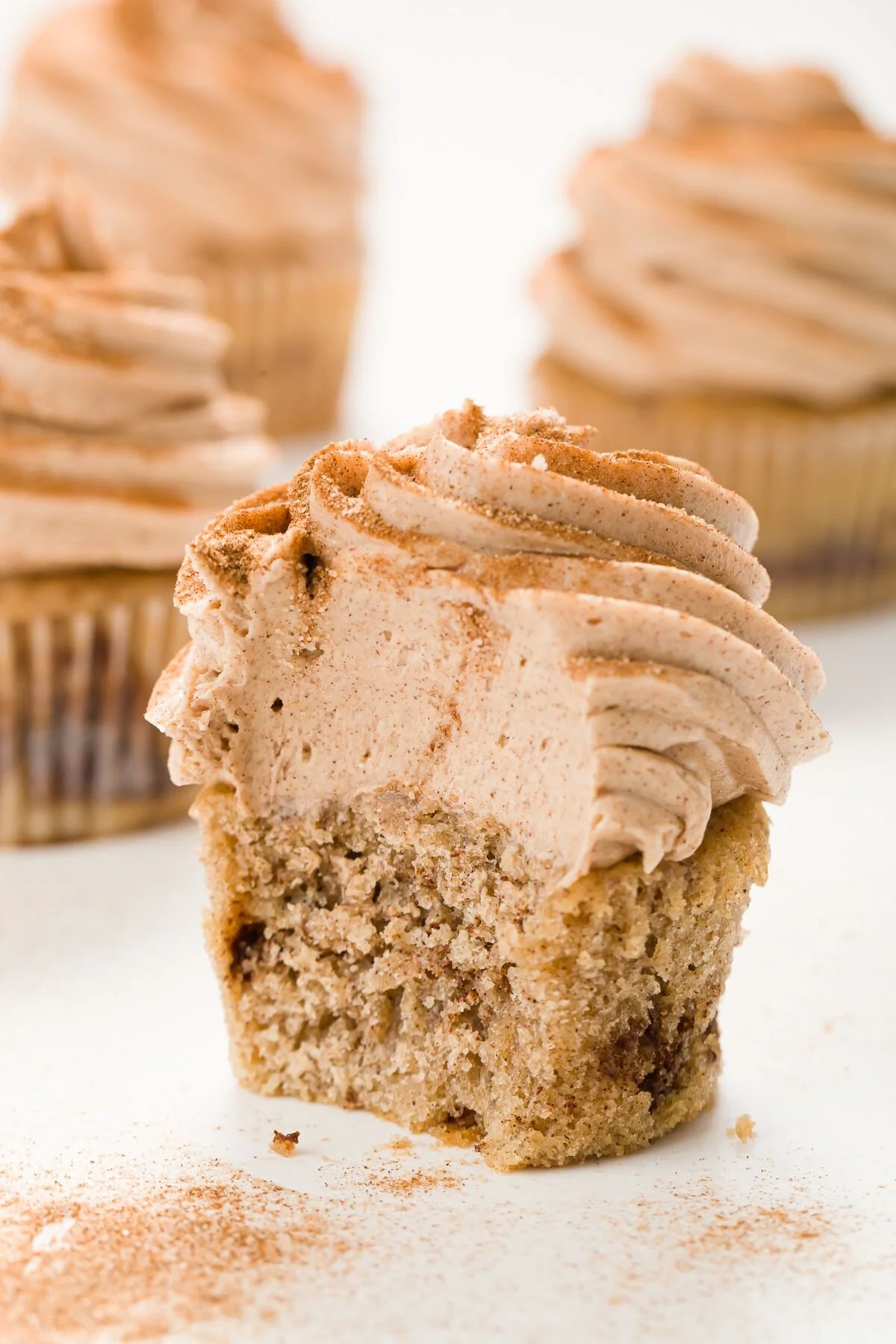 Close-up of a single cupcake with beige frosting topped with cinnamon, in front of two other cupcakes with similar frosting in the background.