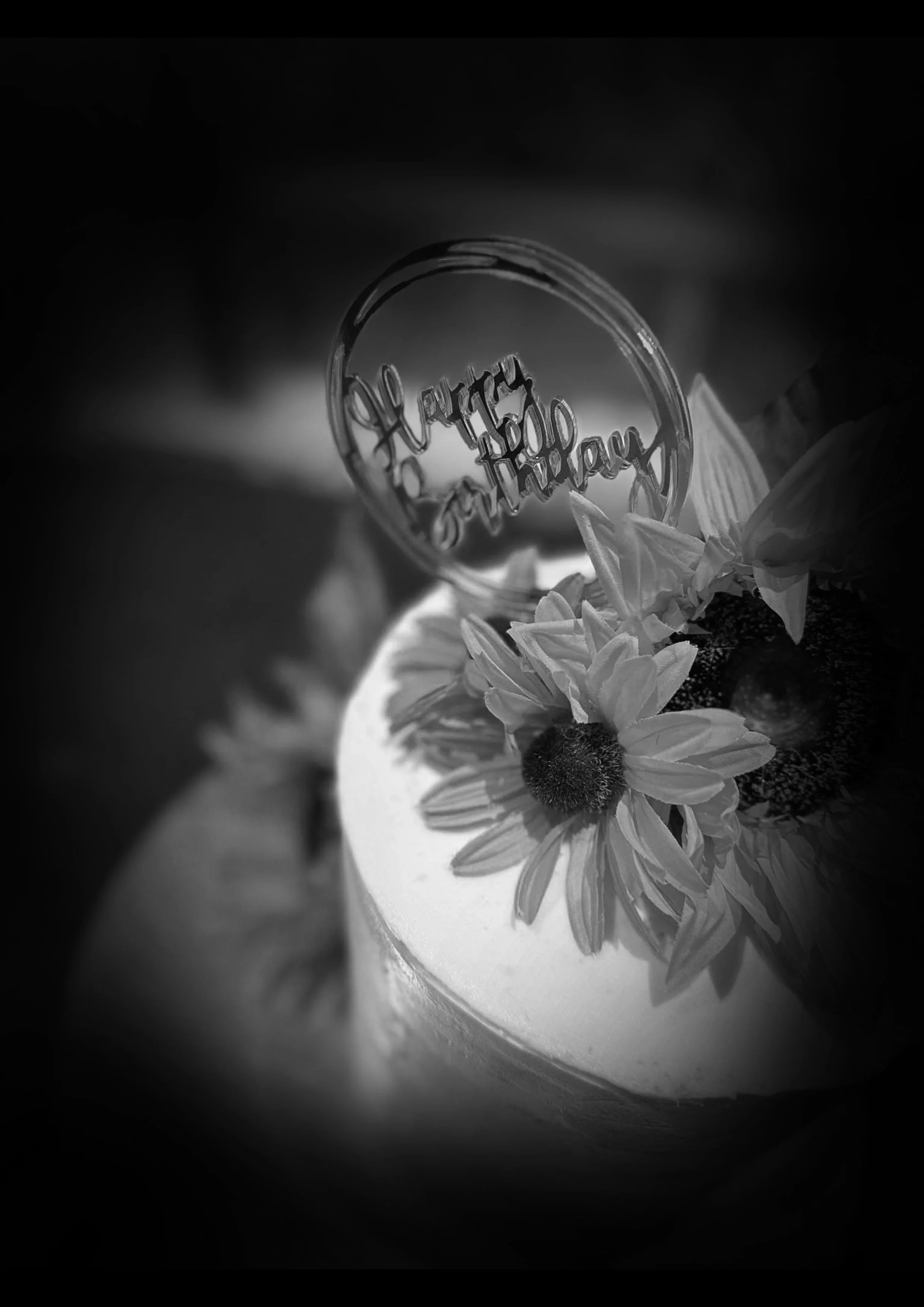 A black and white image of a birthday cake with a decorative topper and flowers, with the words 'happy birthday' in the topper.
