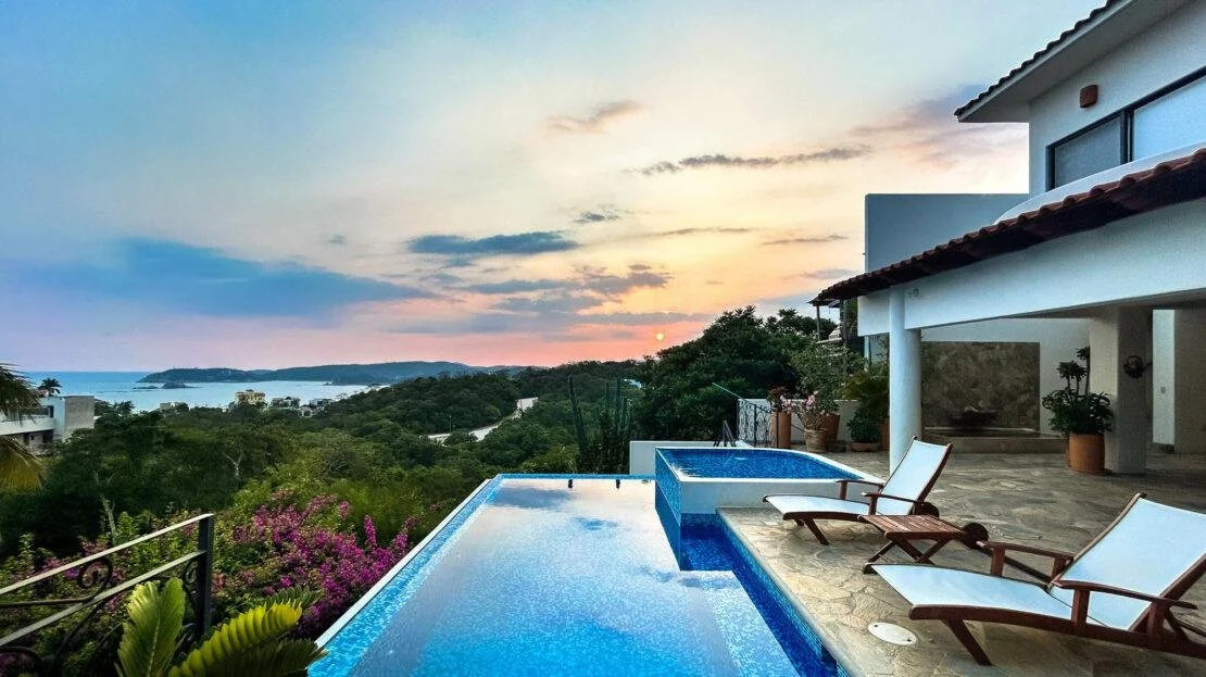 Vista de una alberca infinity con vista al paisaje y el mar, al atardecer, con sillas de descanso junto a la alberca y una casa moderna blanca con techos de teja.