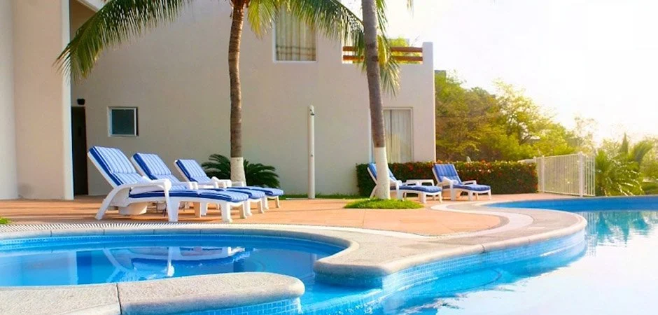 Piscina con tumbonas con cojines azules, palmas y edificio blanco en un ambiente de resort