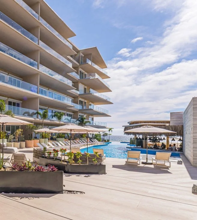 Vista de un complejo residencial con piscina al aire libre y áreas de descanso con sombrillas, en un ambiente soleado junto al mar.