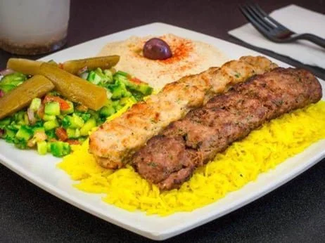 Plate with two grilled kebabs, yellow rice, salad with green vegetables, and a dollop of sauce with a cherry on top.