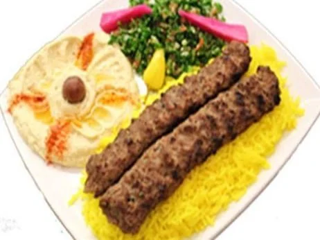 Plate with two grilled kebabs, yellow rice, a salad with greens, pickled turnips, and hummus garnished with a black olive.
