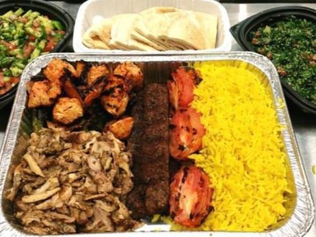 A large meal tray with grilled chicken, beef kebabs, yellow rice, and grilled vegetables, along with side dishes of salad, pita bread, and greens.