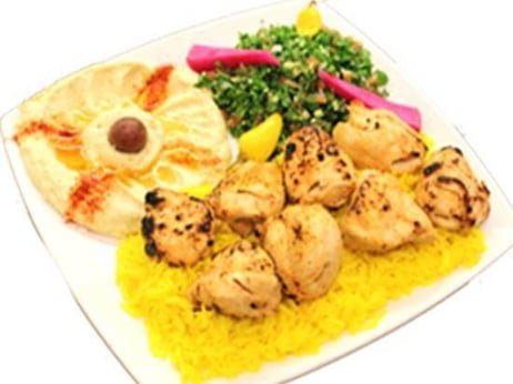 Plate of yellow rice, grilled chicken pieces, green salad with pink radish slices, lemon wedge, and a side of flatbread with a cream-colored sauce and a black olive.