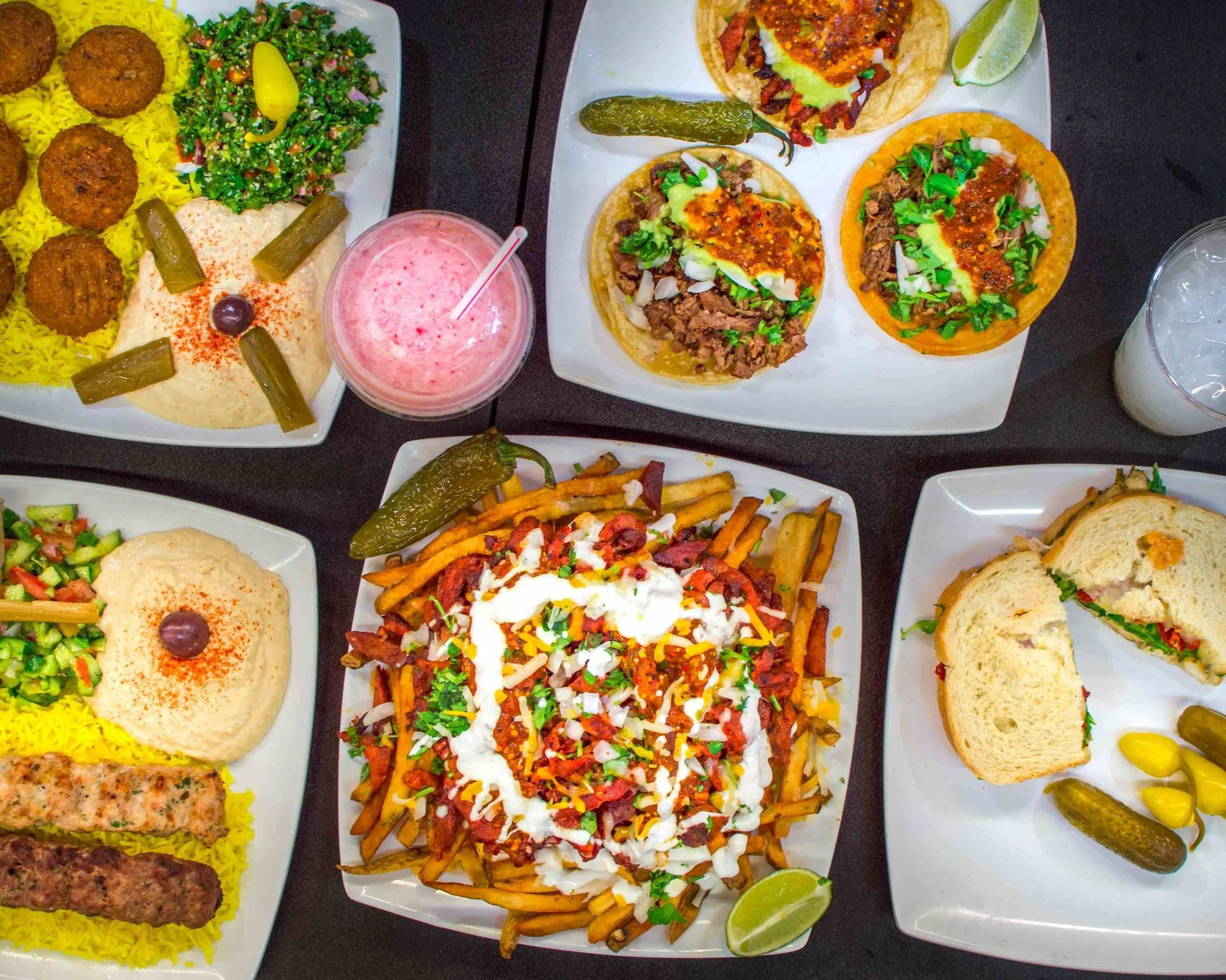 Assorted Mexican dishes including falafel with rice, salad, hummus, and pickles on a white platter, and tacos with meat, onions, cilantro, and sauces on another white platter with lime wedges, chili peppers, and fried potatoes.