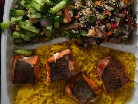 Plate with four pieces of cooked fish on yellow rice, a side of green salad with cucumbers, and a serving of mixed fruit salad with berries and chopped fruit.
