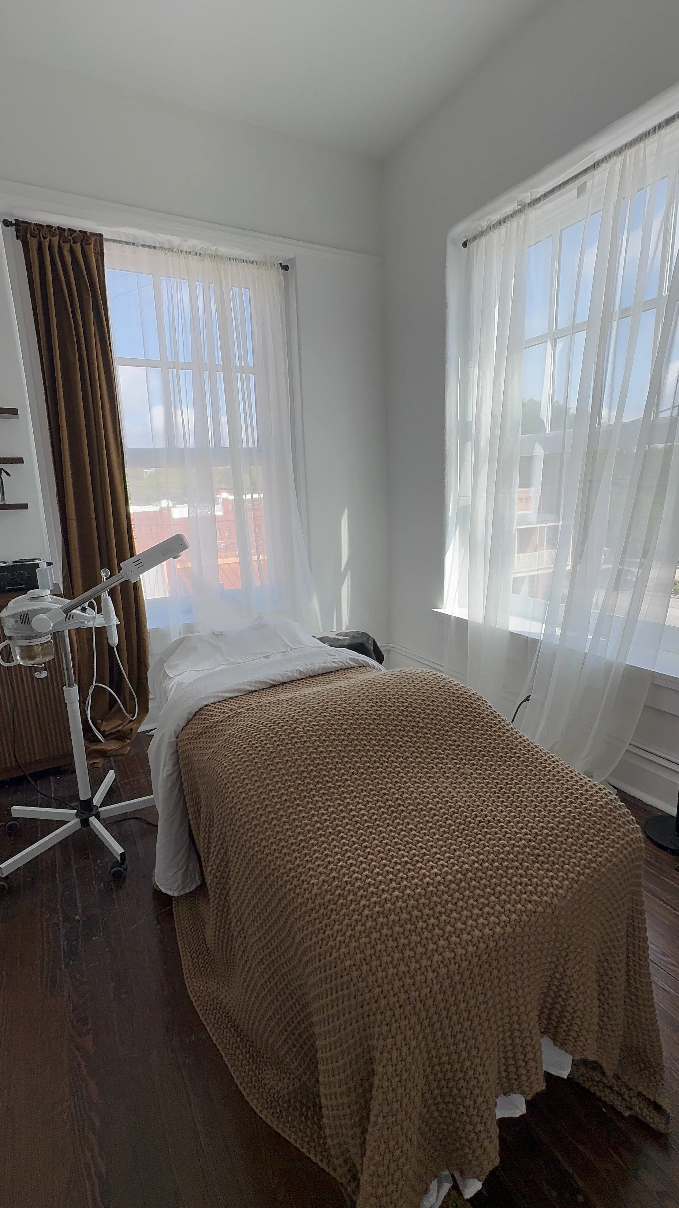 A bedroom with a bed covered by a textured brown blanket, white pillows, near two large windows with sheer white curtains and a brown curtain on one window, natural light coming in, and medical equipment beside the bed.