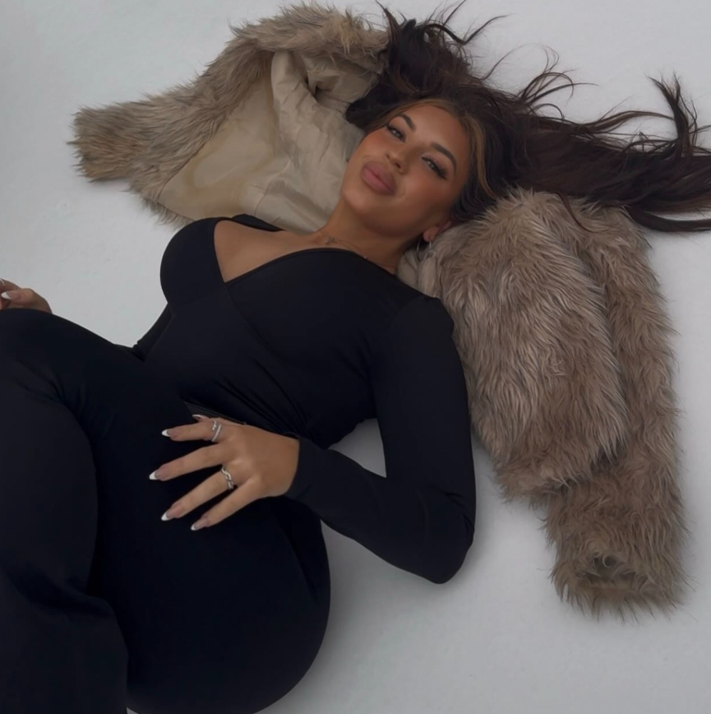 A woman lying on a white surface, resting her head on a fur coat with her hair spread out. She is wearing a black dress with a sheer neckline, and has multiple rings on her fingers. The photo captures her smiling slightly at the camera.