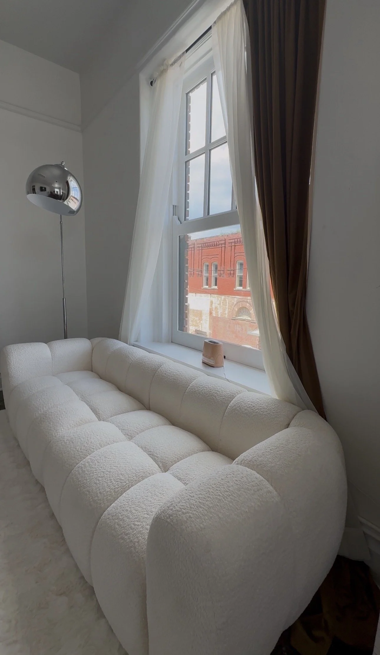 A cream-colored tufted sofa in front of a large window with white curtains, brown drapes, and a view of a red brick building outside. A small pink vase is on the window sill, and a chrome floor lamp stands beside the sofa.