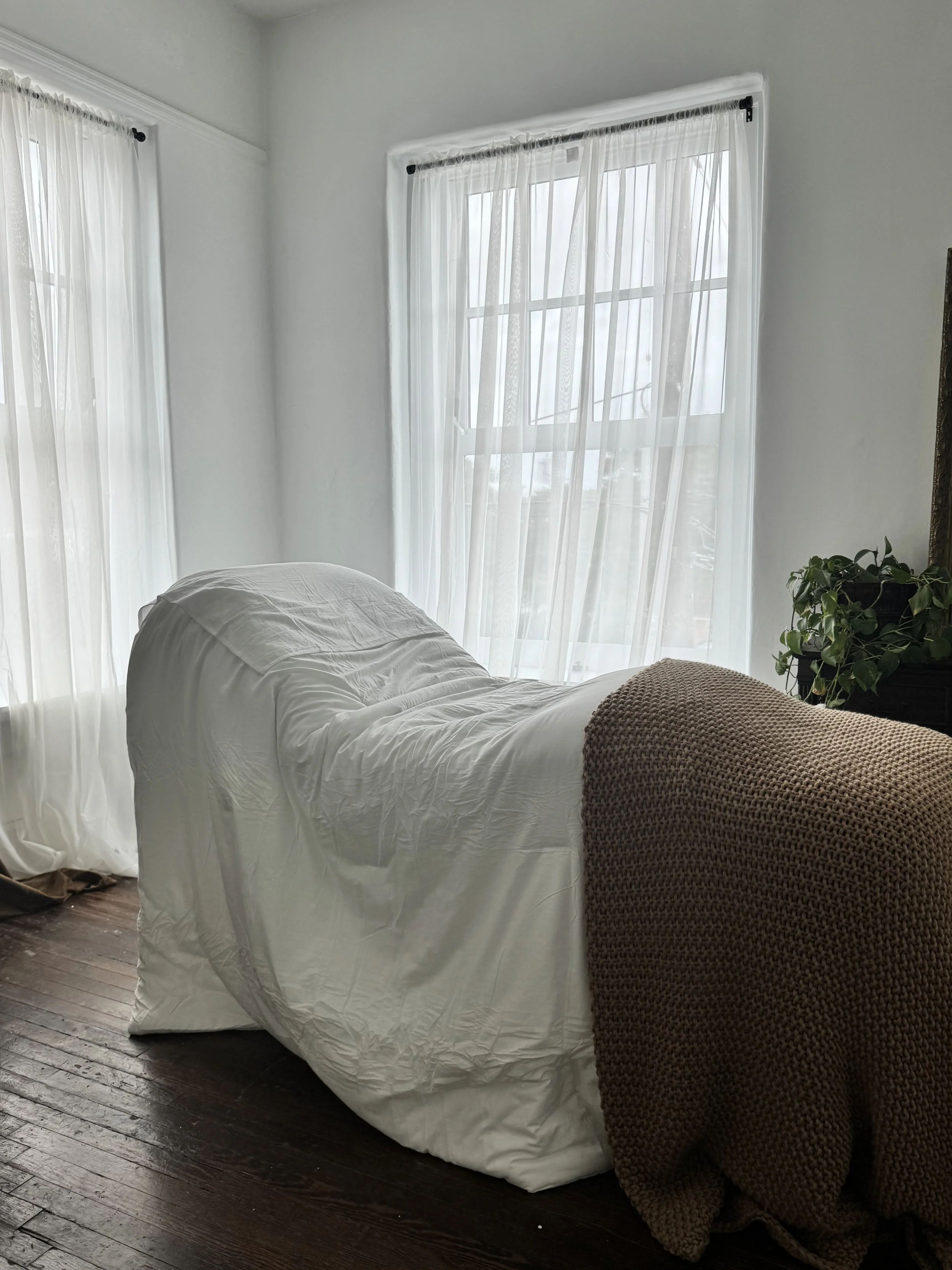 A bed with white bedding and a knotted beige blanket, next to a green potted plant, in a well-lit room with large windows and sheer curtains.
