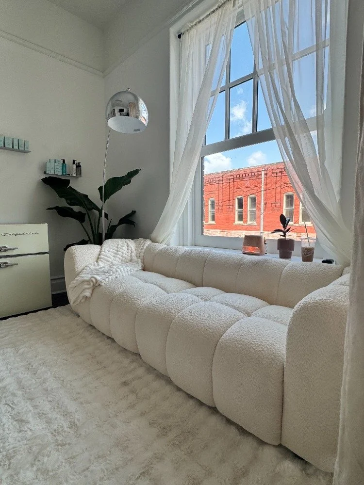 Bright living room with a large white tufted sofa, sheer white curtains, a window overlooking a red brick building, and decorative items on the window ledge.