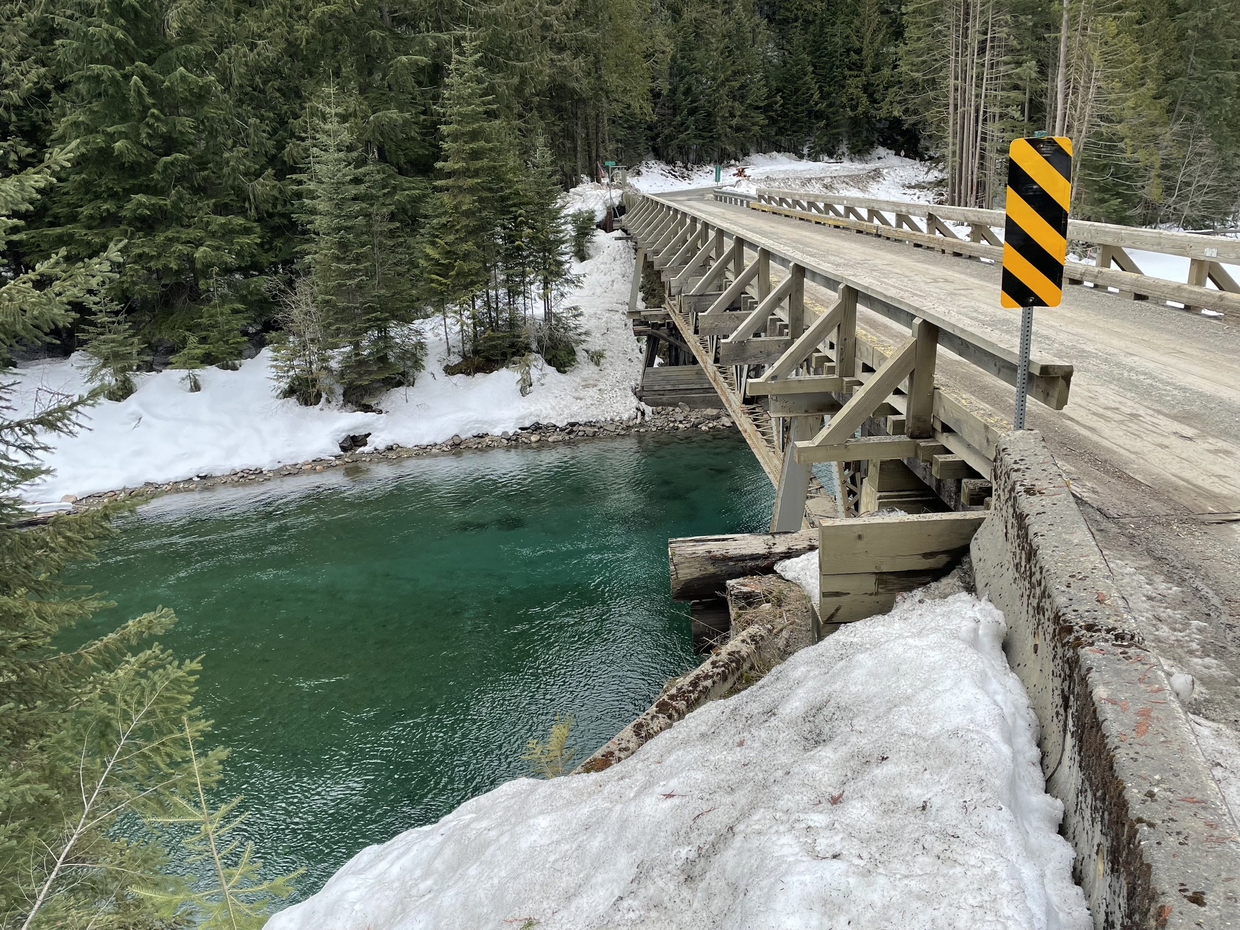 A wooden bridge over a river in a forested, snowy landscape. There is a black and yellow striped warning sign on the right side of the bridge.
