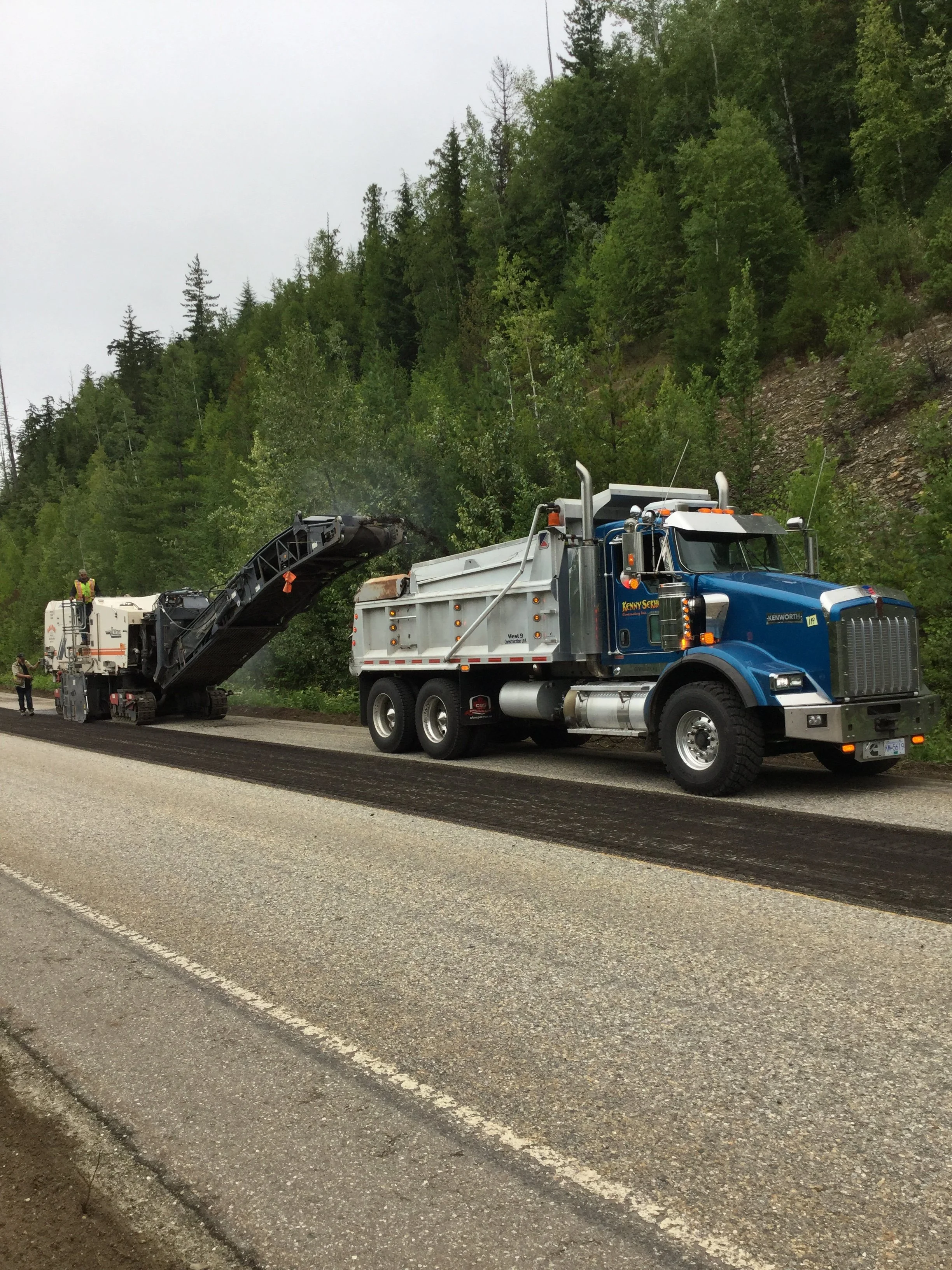 A blue and white road paving truck with workers nearby, laying asphalt on a road in a forested area.