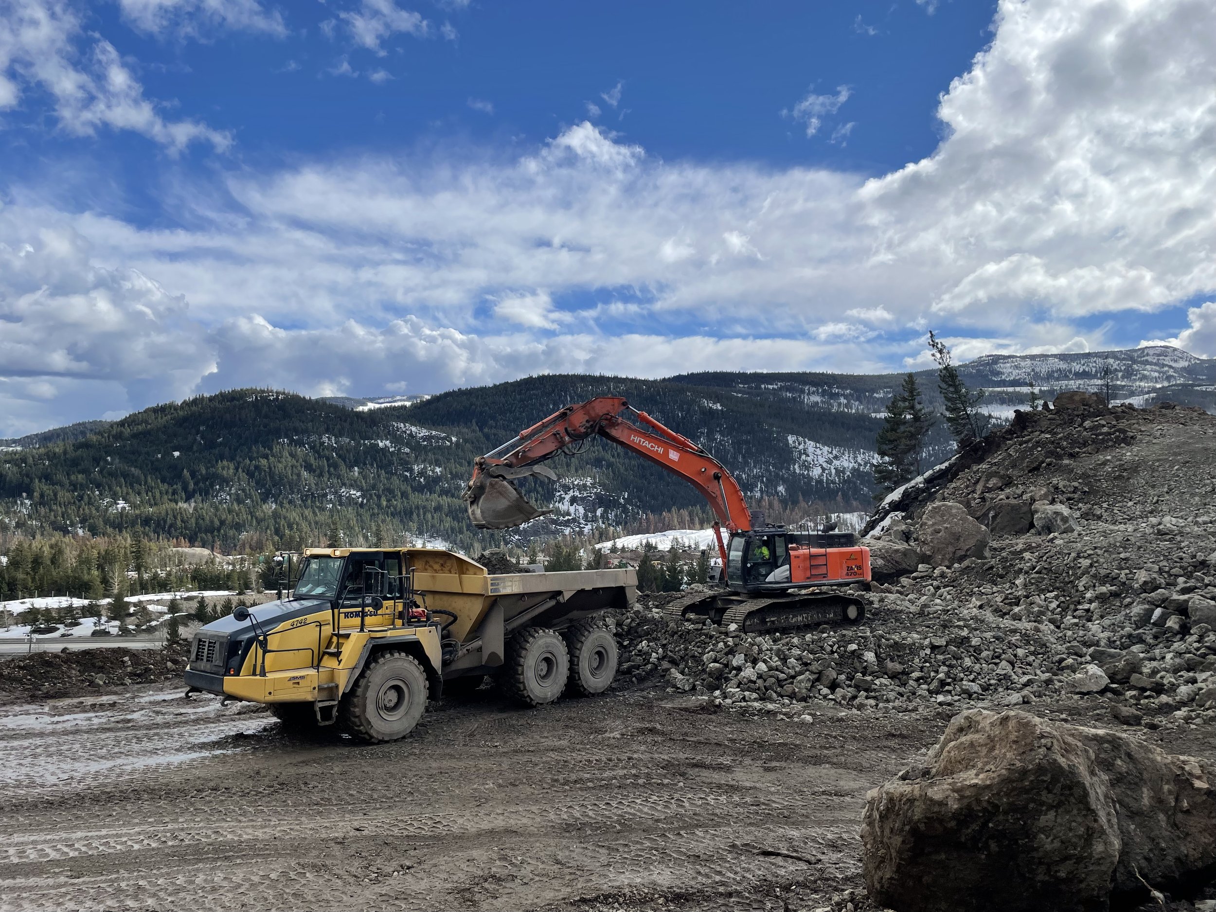 Construction site with large rocks and dirt being moved by an orange Hitachi excavator, a yellow Komatsu bulldozer, and a dump truck, set against a backdrop of forested mountains and a partly cloudy sky.