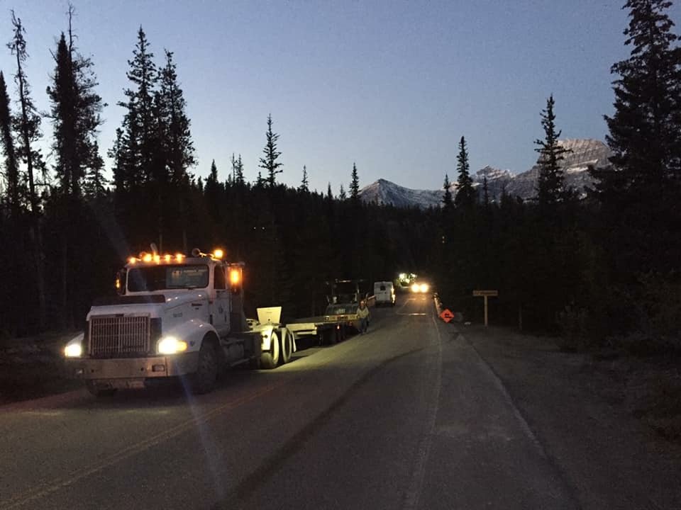 A large truck is parked on the left side of a rural road during dusk, with several smaller vehicles behind it. The scene is surrounded by dense pine trees and mountains in the background, with the sky darkening.
