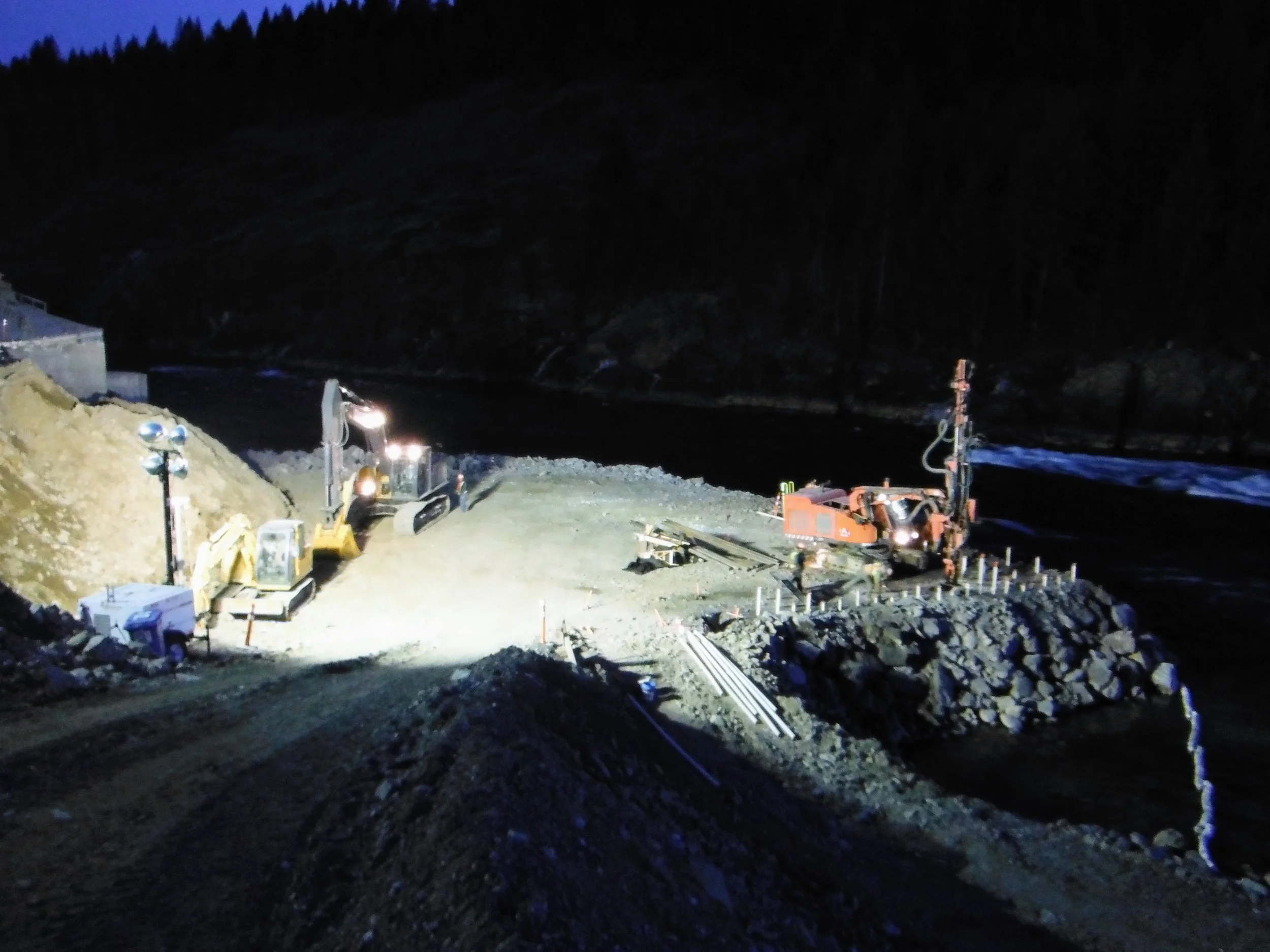 Nighttime view of a construction site on a hillside with equipment, lights, and materials scattered around. The background shows a dark, forested mountain.