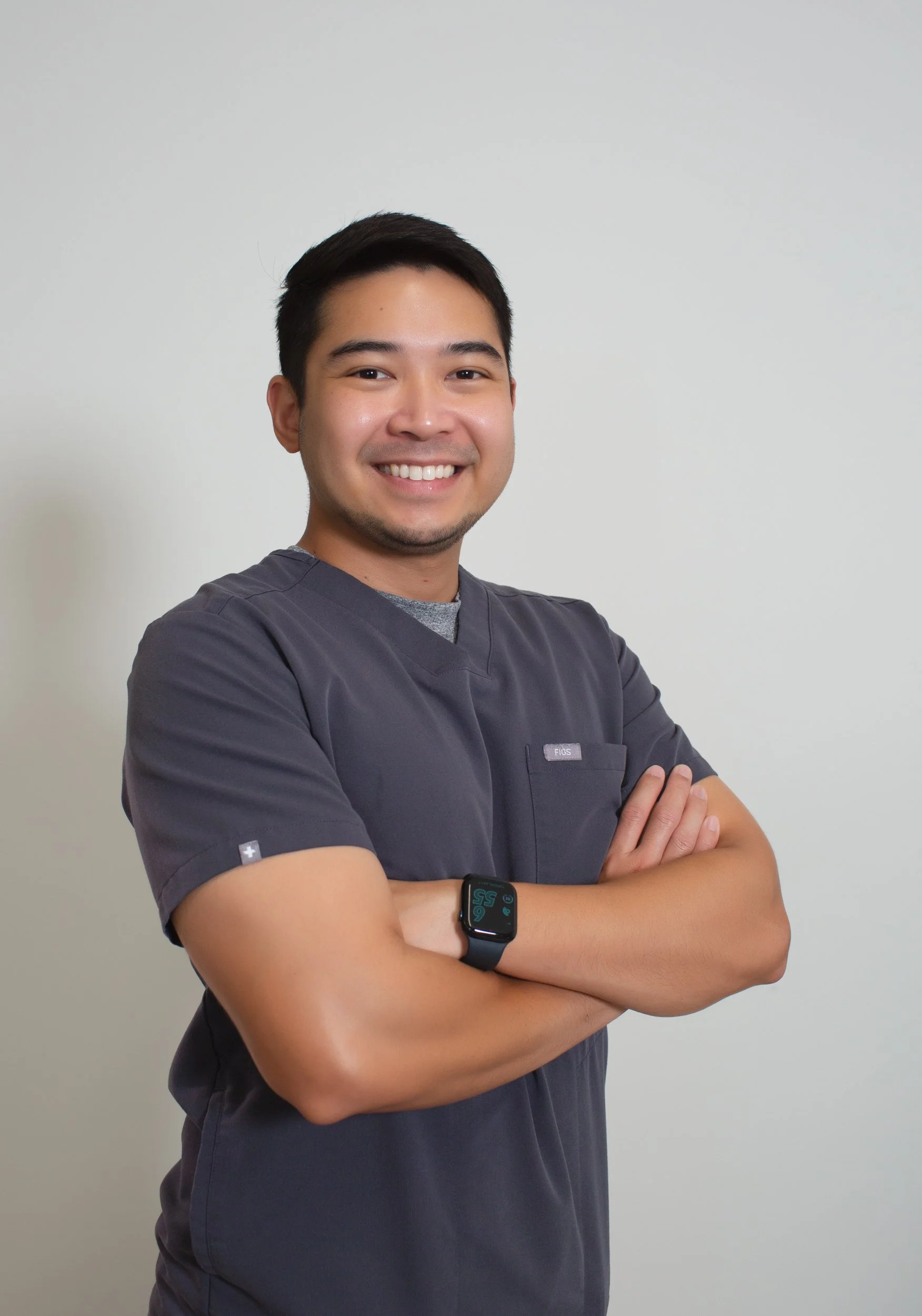 A smiling man wearing medical scrubs with arms crossed, standing against a plain white background.