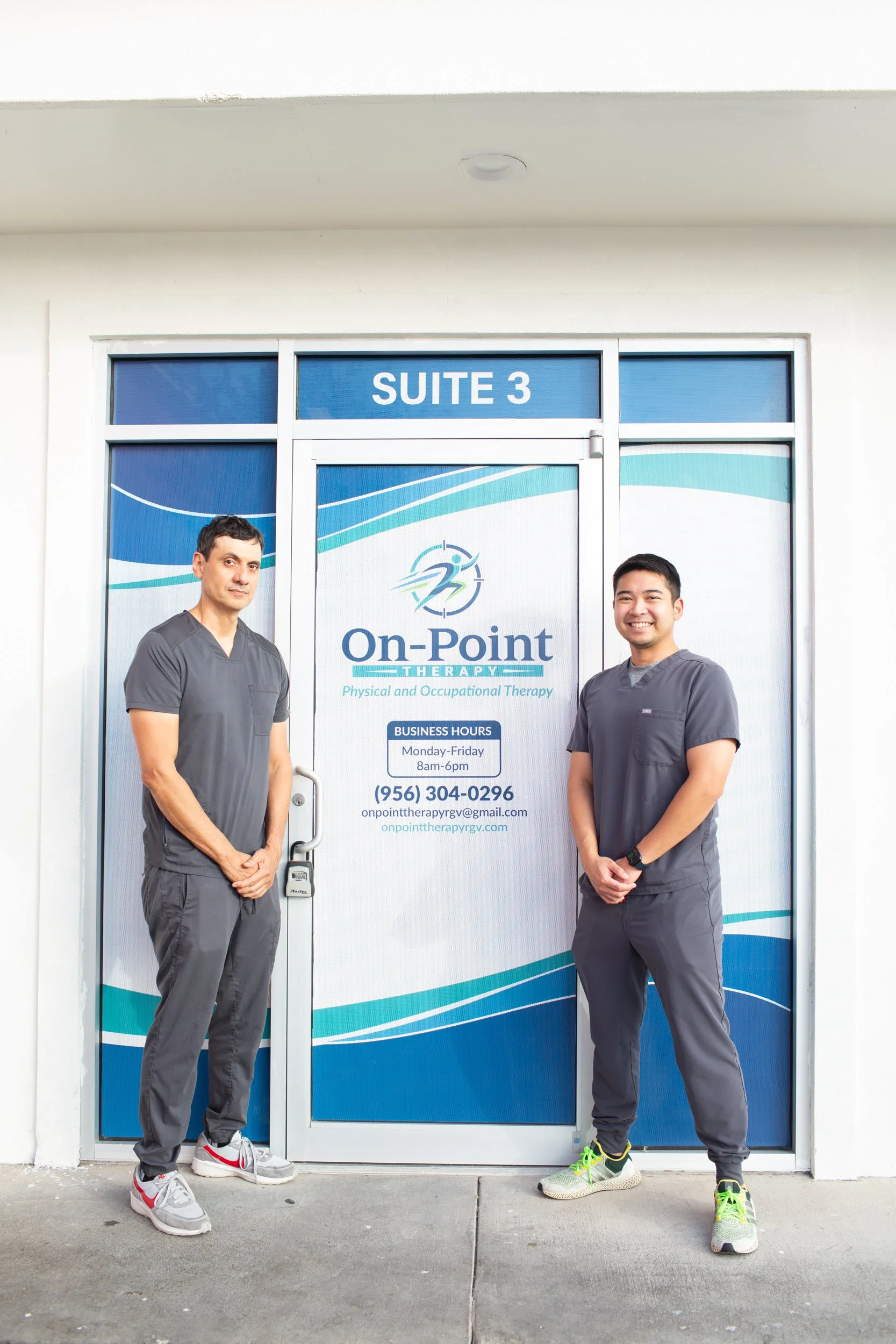 Two men in gray medical scrubs standing outside the entrance of On Point Therapy, a physical and occupational therapy clinic, in front of a glass door with the clinic's sign, suite 3, business hours, phone number, and email address.