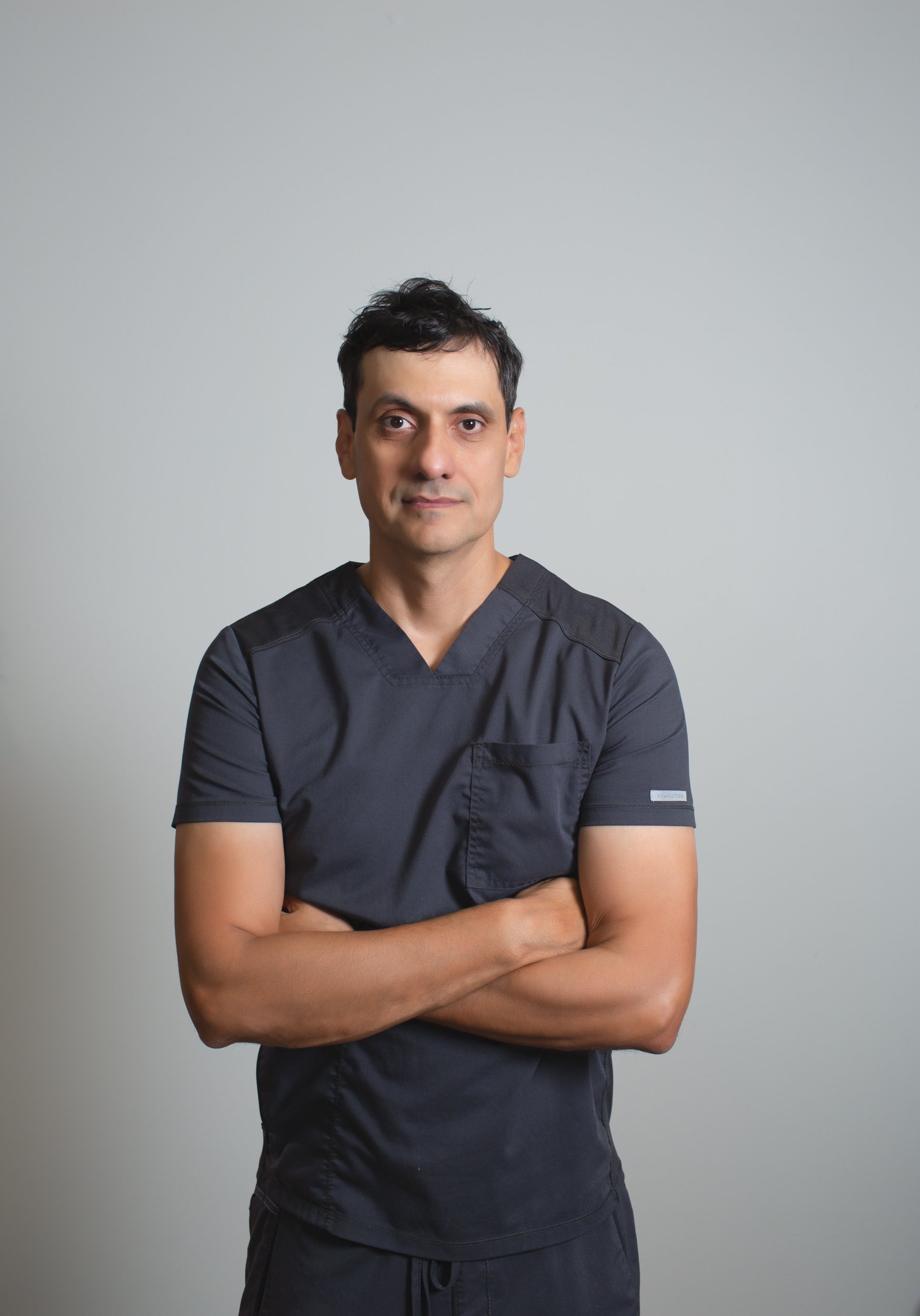 A man with dark hair and light skin wearing dark scrubs, standing with arms crossed against a plain light gray background.