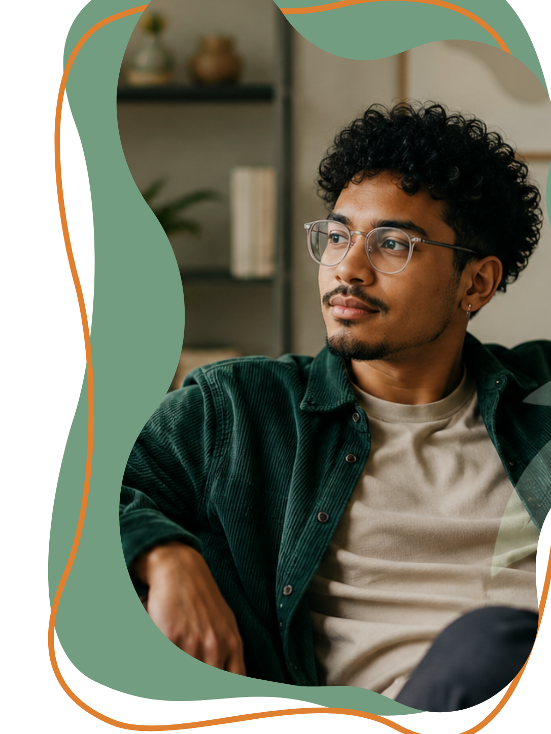 A young man with curly hair and glasses sitting in a room with a bookshelf in the background. He is wearing a green shirt over a beige t-shirt and appears to be relaxed, looking thoughtfully to the side.