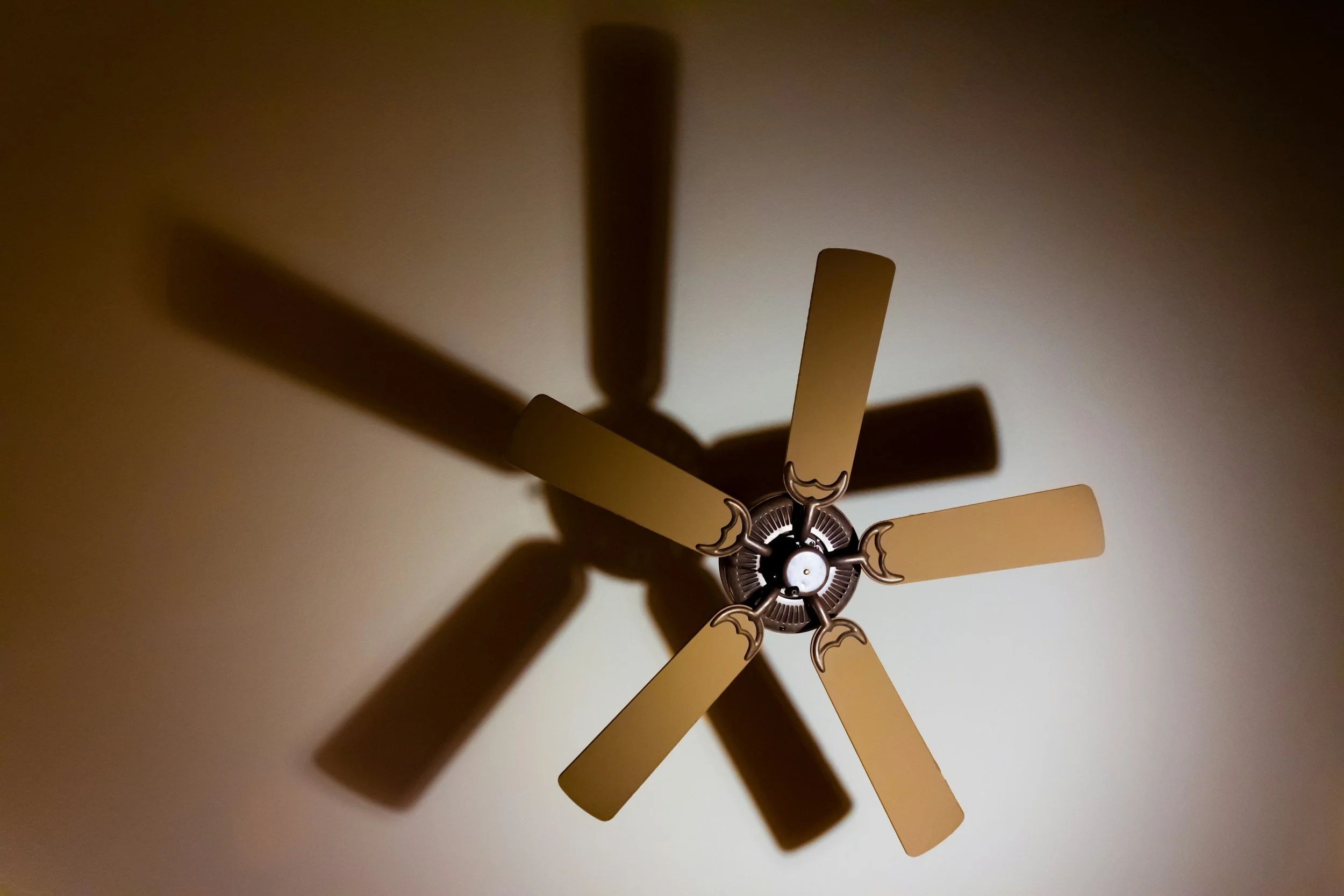 Close-up of a ceiling fan with five beige blades and decorative metal accents, casting shadows on the ceiling.