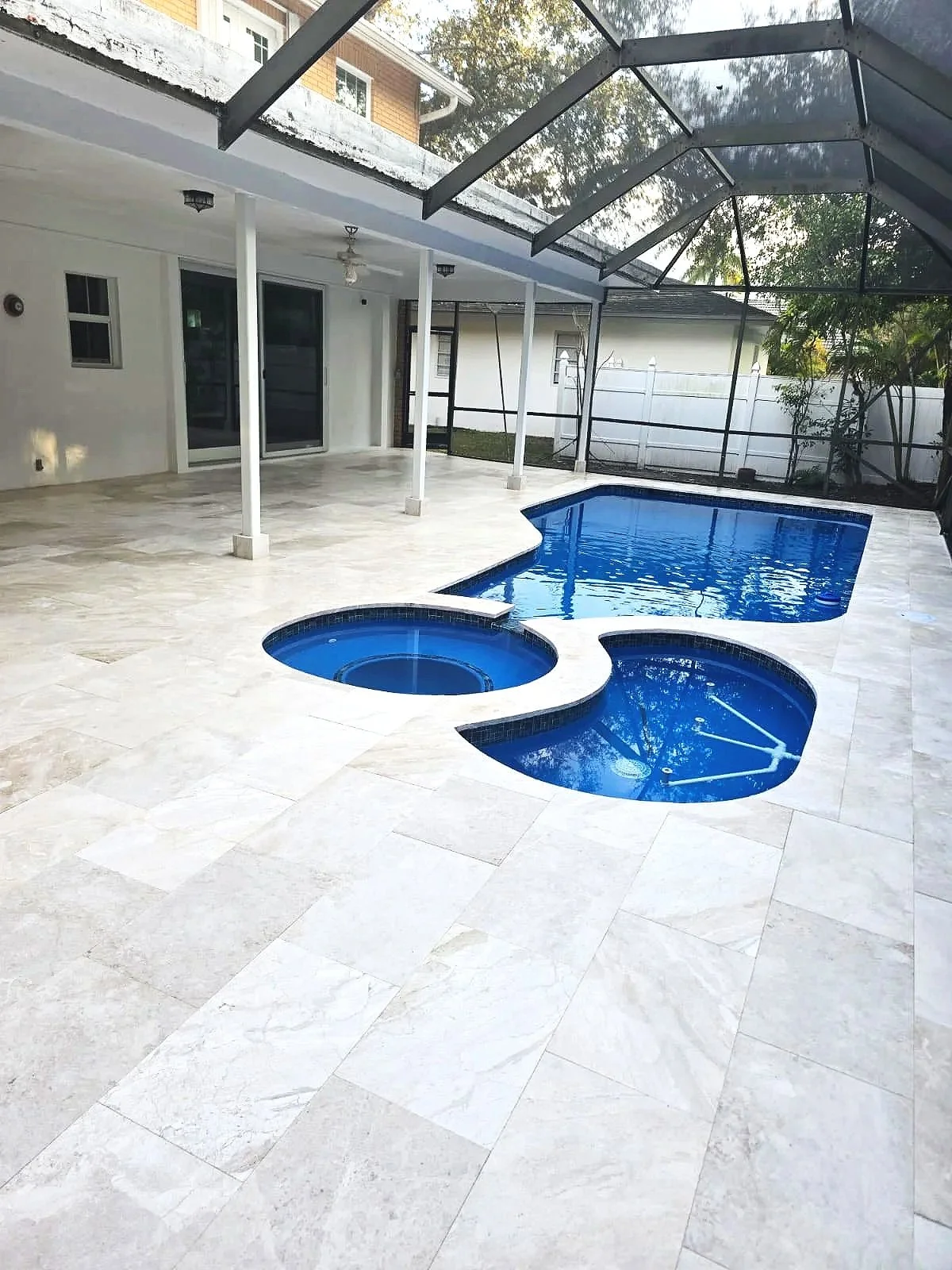 A backyard enclosed patio with a screened pool and hot tub surrounded by beige tile flooring, white walls, sliding glass doors, outdoor ceiling fan, and a black metal and screen enclosure.