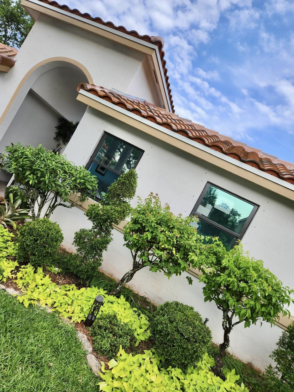 A white house with a red tile roof and large rectangular windows, surrounded by a well-maintained garden with various green bushes and trees, under a partly cloudy blue sky.