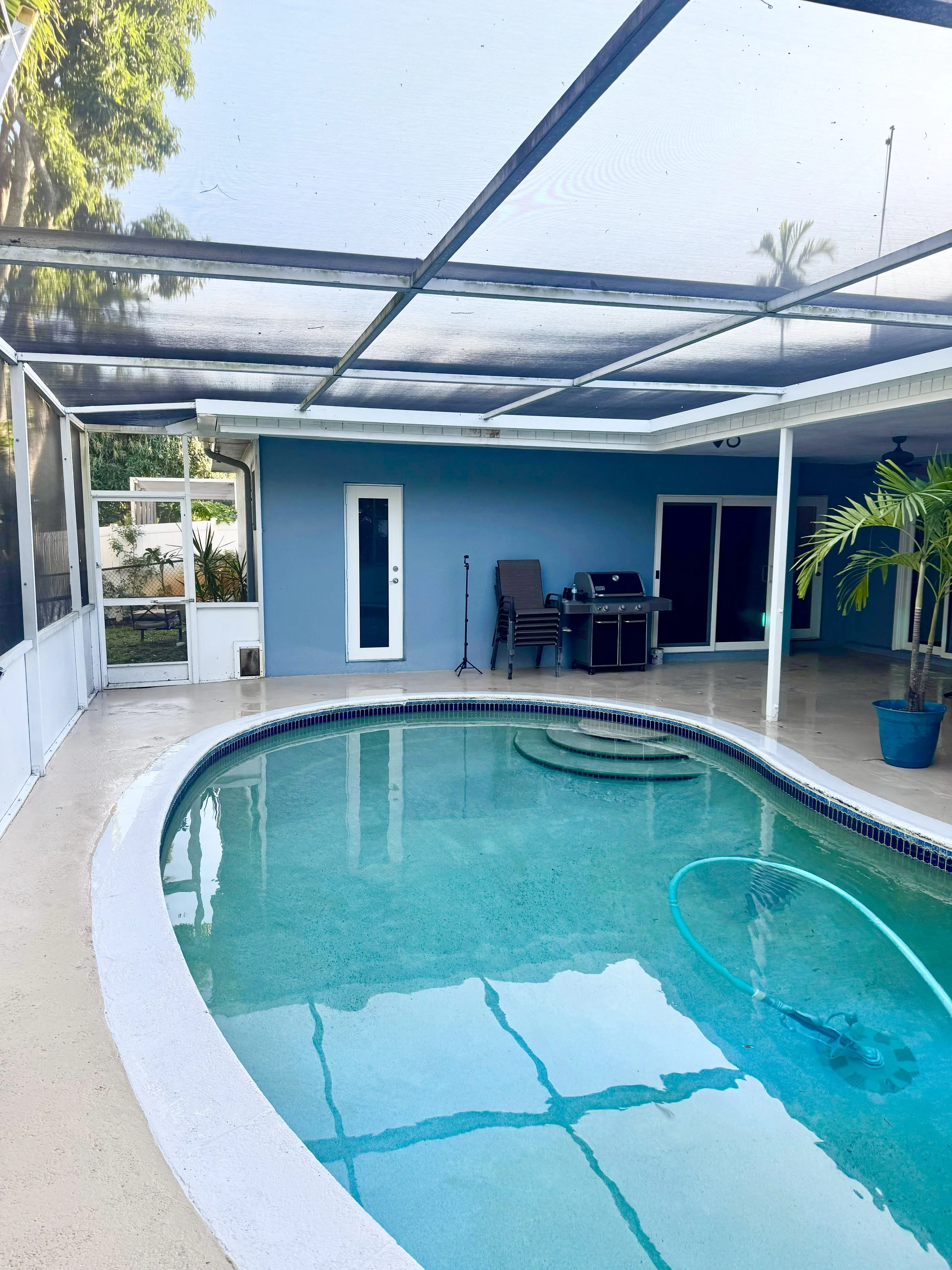 In-ground backyard swimming pool with a pool vacuum in the water, surrounded by a concrete deck that was pressure washed by sian home services in broward county, with a blue house wall, and a screened porch area renovated by Sian home services.