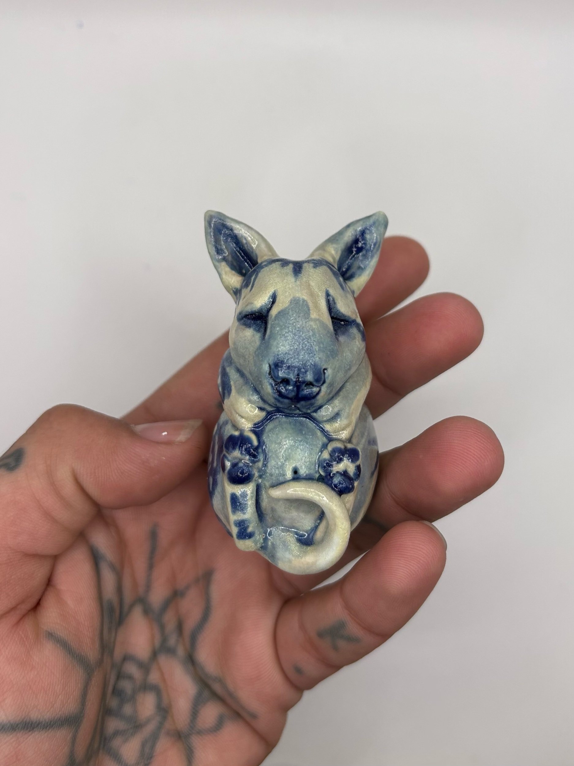 A person holding a ceramic or clay figurine of an animal, possibly a fox or dog, with a white and blue pattern, against a plain white background.