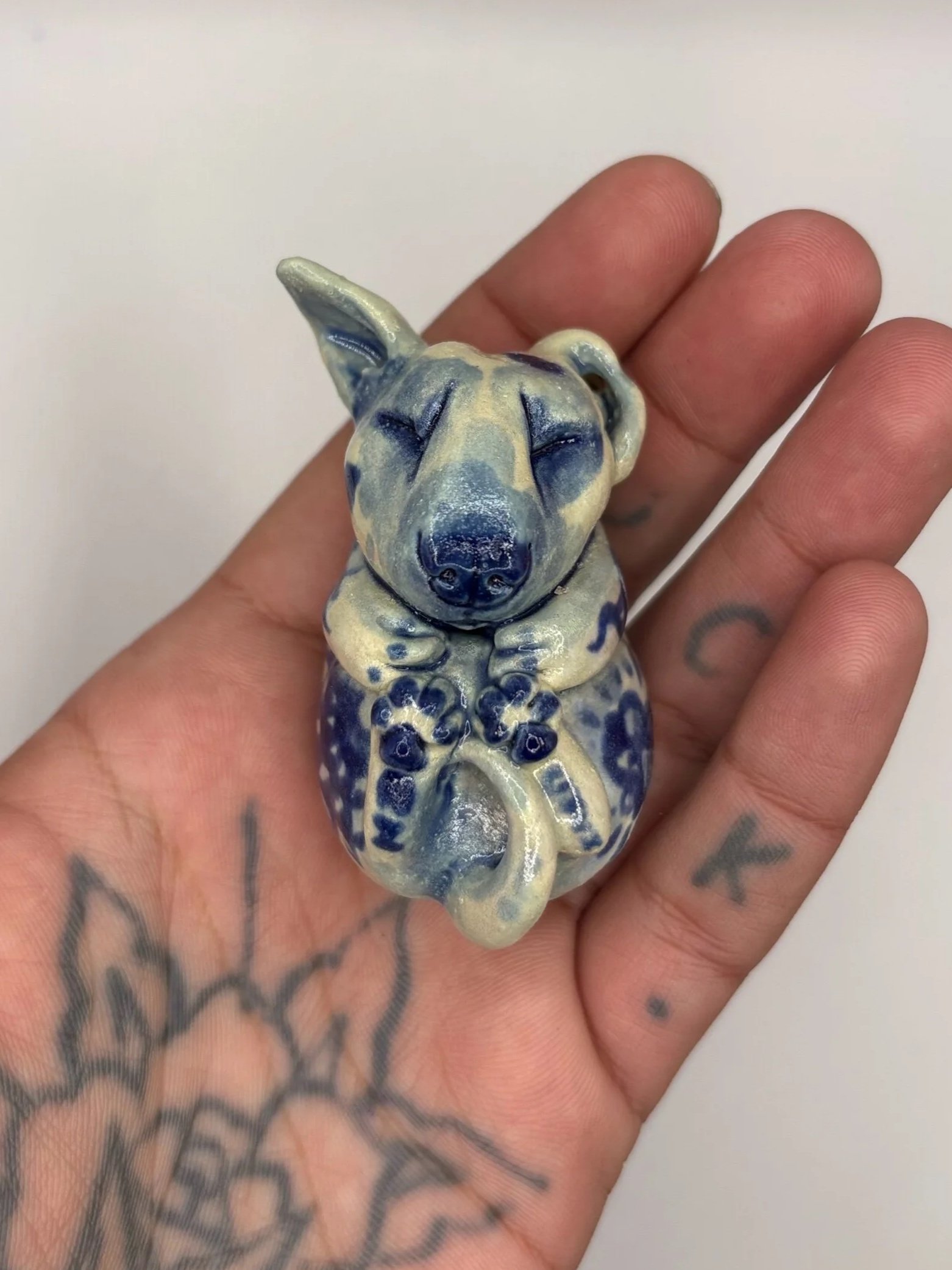 A person holding a ceramic sculpture of a dog with blue and cream colors. The person has tattoos on their hand and fingers, and a floral tattoo on their wrist. The background is plain and light-colored.