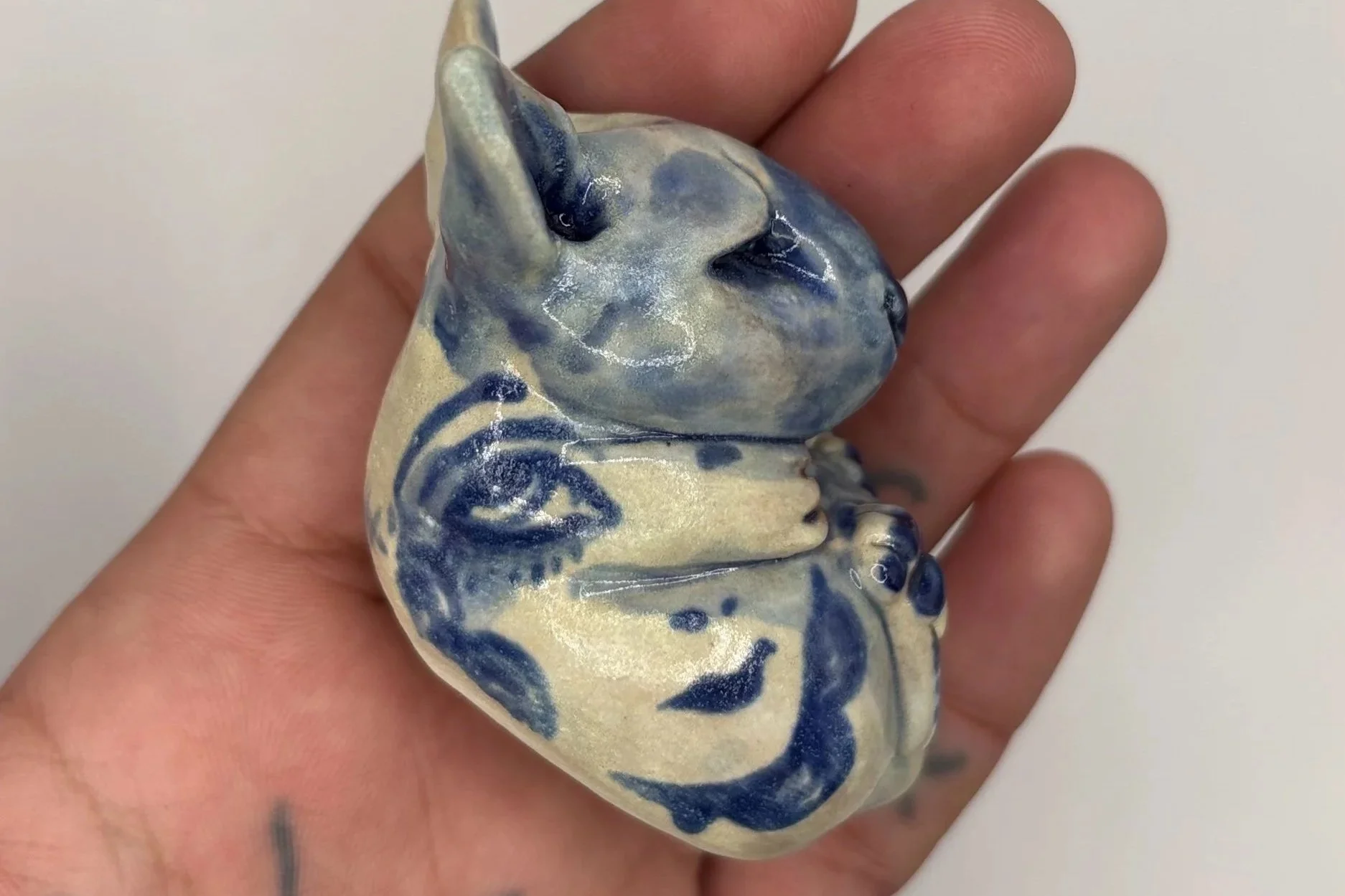A person's hand holding a small ceramic figurine of a baby or child with a cat's face painted on it, featuring blue and white glaze details.