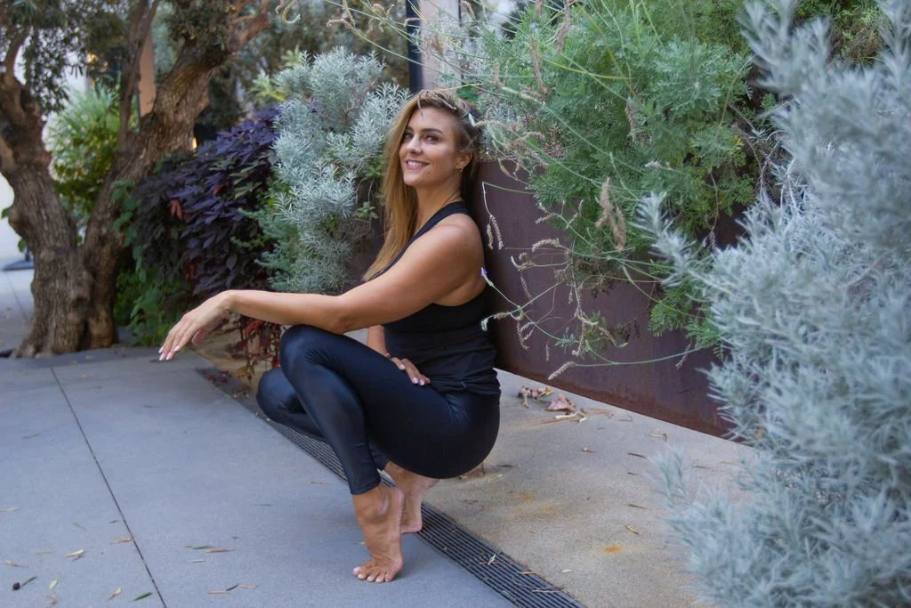 Nicole Sciacca of Sweat + Stillness in a black tank top and leggings squats on a sidewalk next to green and gray plants, smiling at the camera.