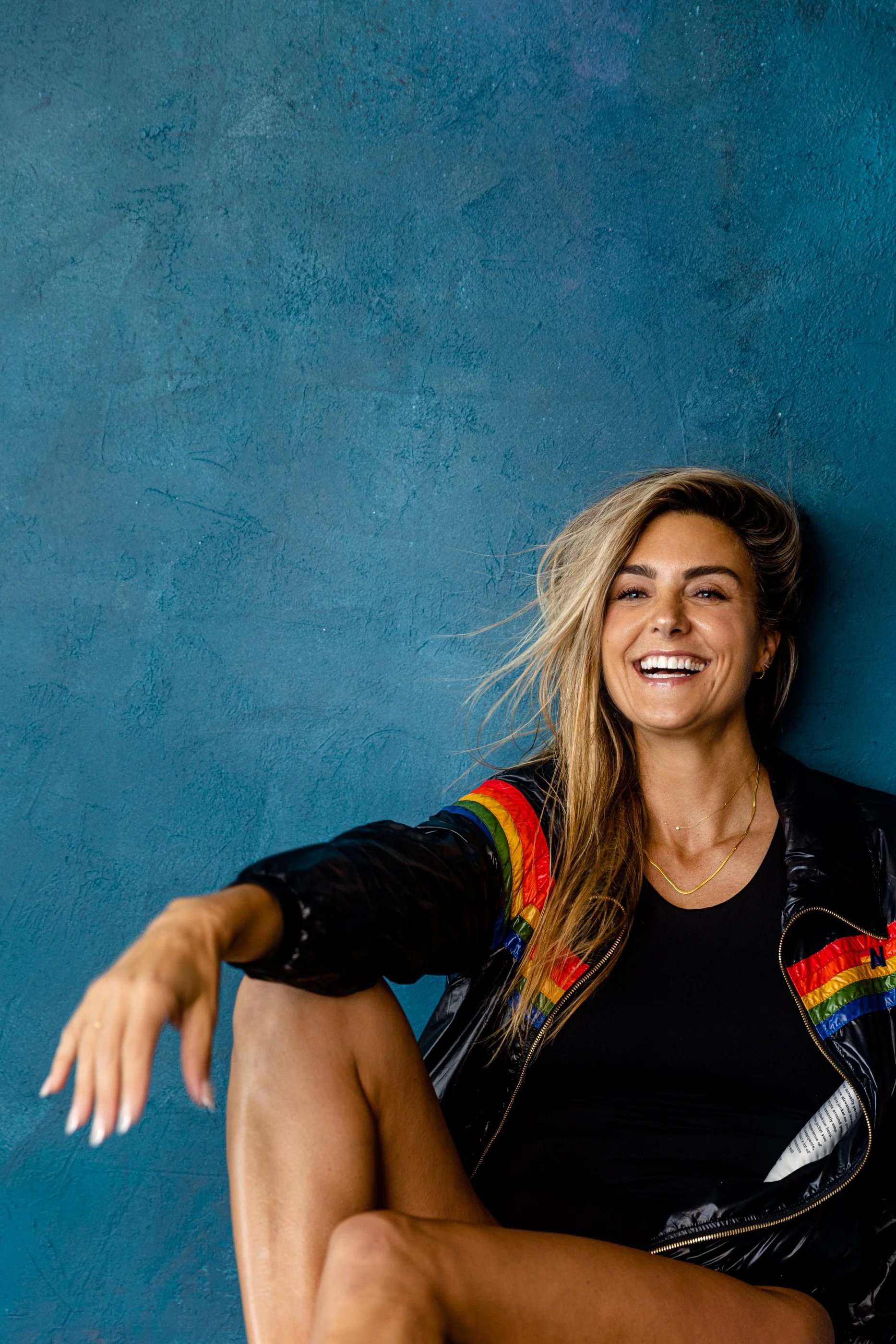 Sweat + Stillness founder Nicole Sciacca with long blonde hair smiling and taking a selfie against a textured blue wall. She is wearing a black top and a rainbow-colored jacket with a zipper.