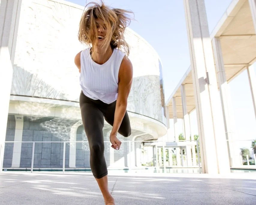 Nicole Sciacca of Sweat + Stillness working out with messy hair in workout clothes performs a one-legged balance pose outside a modern building.