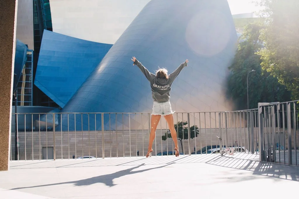 Photo of Nicole Sciacca of Sweat + Stillness with arms raised in front of the Walt Disney Concert Hall, with sunlight shining and a small scooter on the ground.
