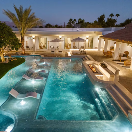 Retreat Photo: Luxury backyard with illuminated swimming pool, lounge chairs, palm trees, and outdoor seating area at dusk.