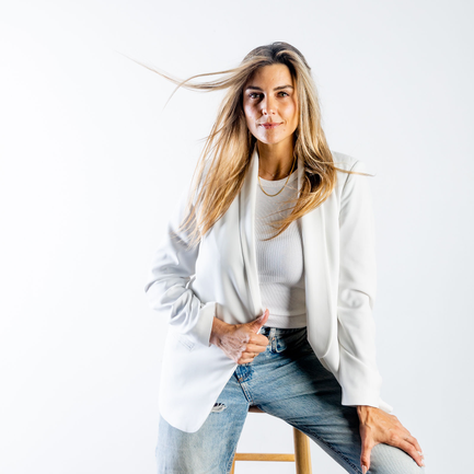 Nicole Sciacca of Sweat + Stillness with long blonde hair wearing a white blazer and jeans, sitting on a stool against a white background.