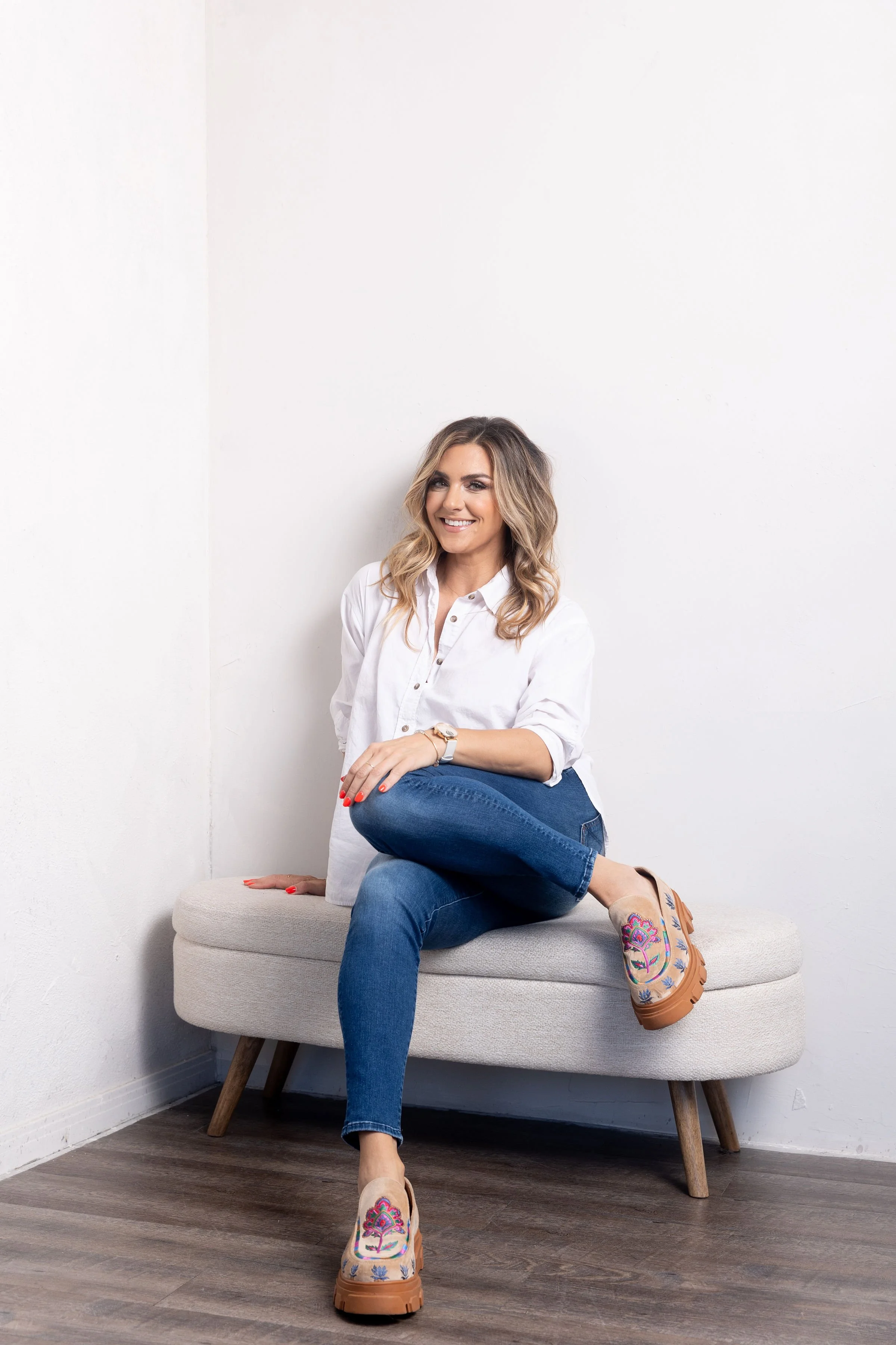 Nicole Sciacca of Sweat + Stillness is sitting on a light gray cushioned bench in a room with white walls and dark wood flooring. She is smiling, with her left leg crossed over her right and her right hand resting on the bench.