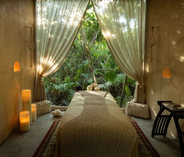 RETREATS: A cozy massage room with a massage table covered in a beige blanket, open to a lush, green outdoor view through large, flowing curtains. Candle lights are placed on the floor near the wall, creating a warm ambiance.