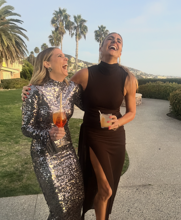 Nicole Sciacca of Sweat + Still laughing and enjoying drinks with friend outdoors with palm trees and a hilly landscape in the background. One woman wears a shiny, sequined dress, and the other wears a black dress with a thigh-high slit.