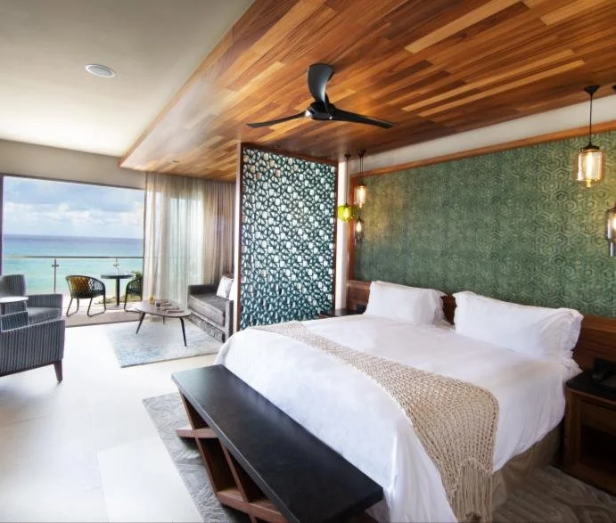 Retreat Photo: Beachside hotel room with a king-sized bed, green patterned accent wall, wooden ceiling, and a balcony with sea view and outdoor seating.