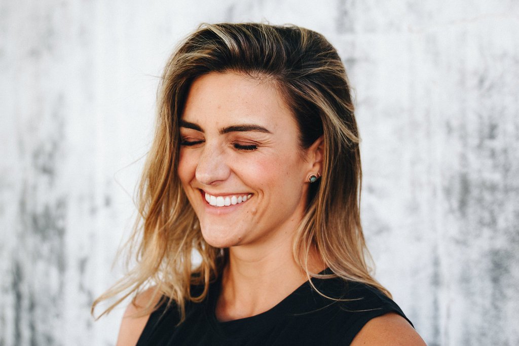 Close-up of a smiling Nicole Sciacca of Sweat + Stillness with wavy blonde hair and earrings, wearing a black top, against a textured white wall background.