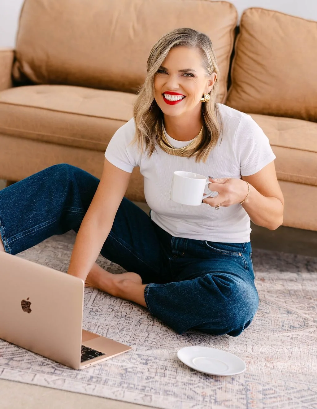 Laura, Nicole Sciacca's retreat co-host sitting on the floor with a laptop in front of her, holding a white mug, smiling, wearing a white t-shirt, gold jewelry, and dark jeans, in a cozy living room.