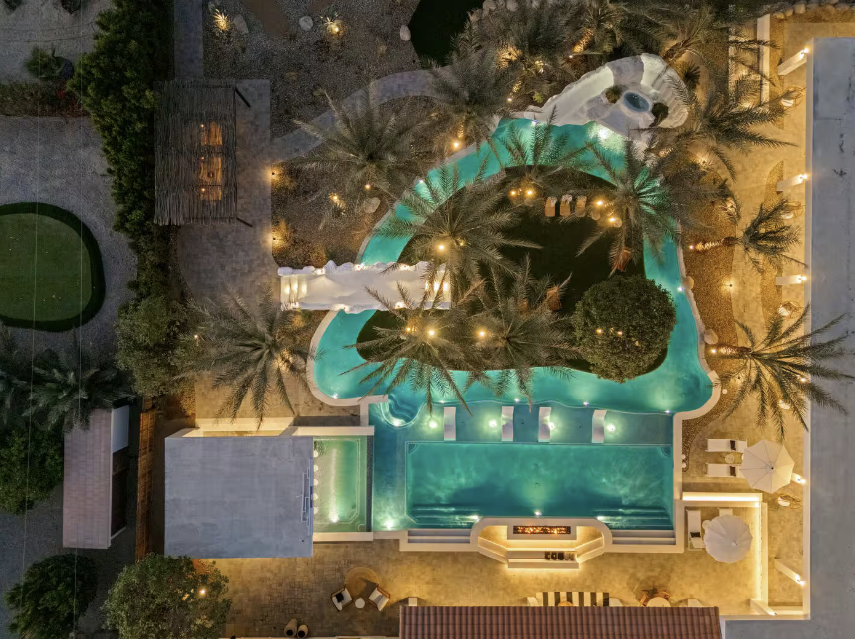 Nighttime aerial view of a swimming pool area with palm trees, outdoor seating, and lighting