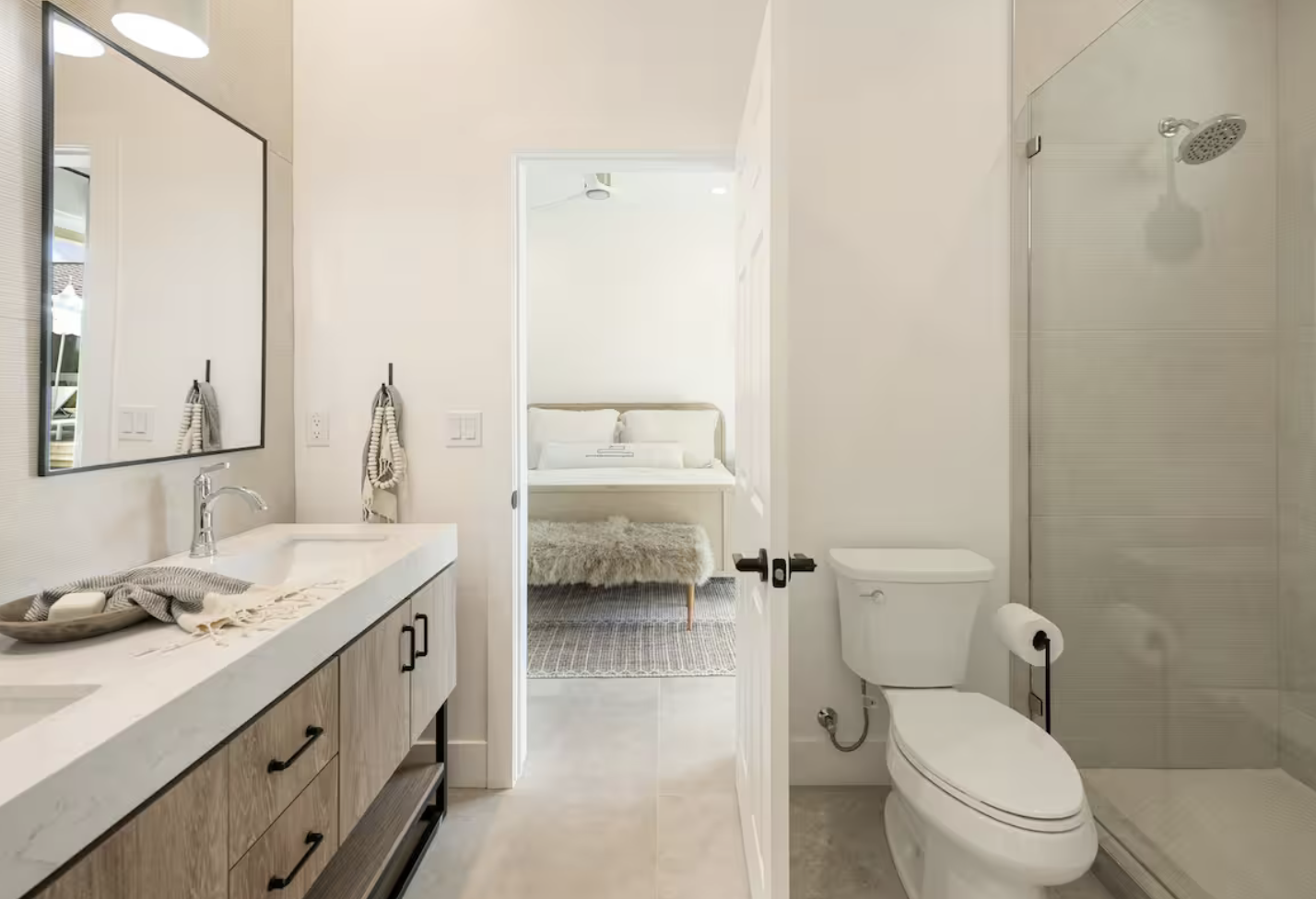 Bathroom with a vanity mirror, sink, towels, and a shower on the right, and a view into a bedroom with a bed and rug.