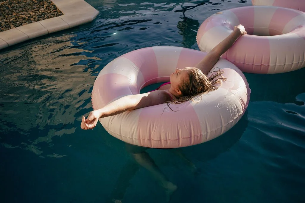 A young woman with wet hair relaxing on a pink and white inflatable pool float in a swimming pool, stretching with arms and legs extended.