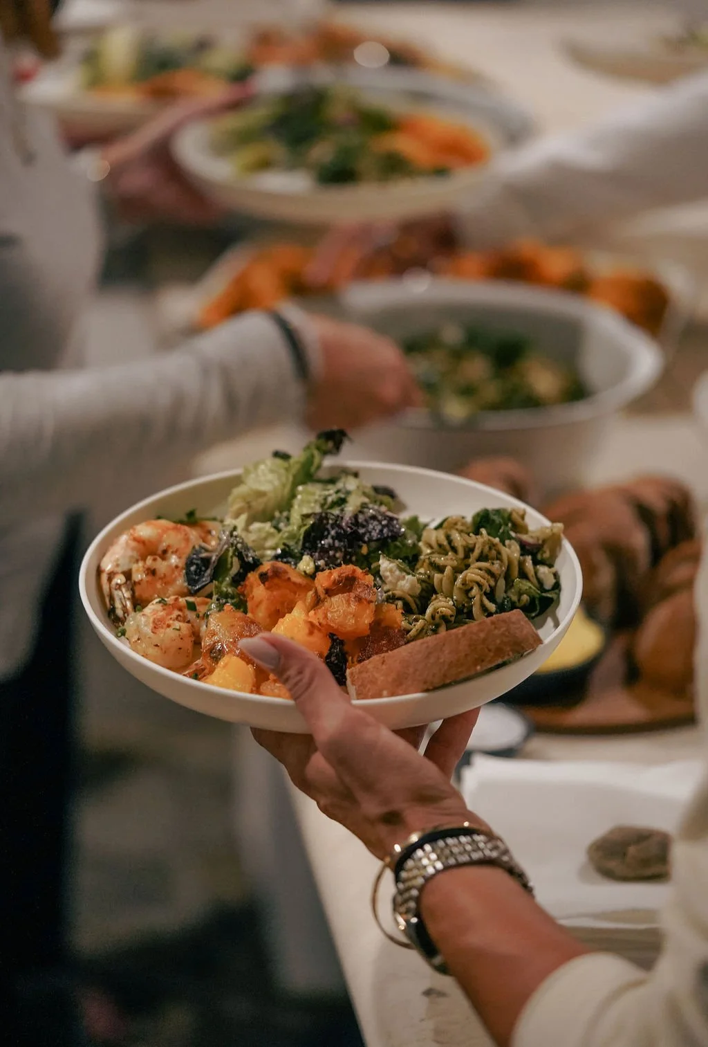A person holding a bowl of food with various side dishes including pasta, vegetables, and bread, at a buffet or gathering with multiple dishes on a table in the background.