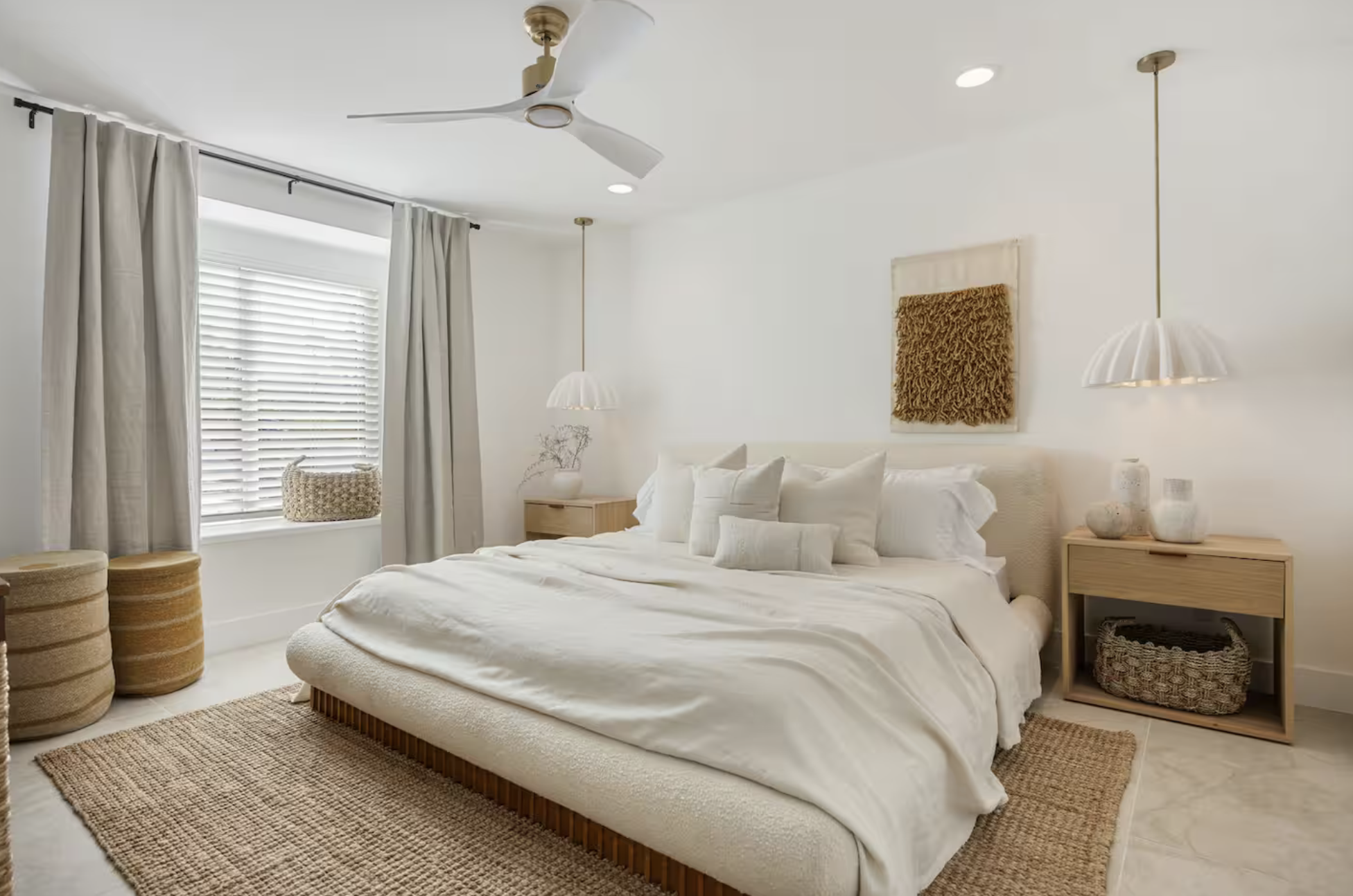 A bedroom with a white bed, beige bedding, and multiple white pillows. Light wooden bedside tables with vases, hanging pendant lights, a window with blinds and beige curtains, a ceiling fan, and woven baskets on a beige rug.