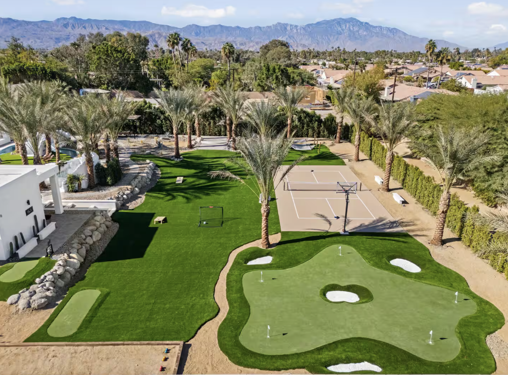 Aerial view of a recreational outdoor area with palm trees, a mini golf course, two sports courts, and a landscaped backyard, with mountains in the background.